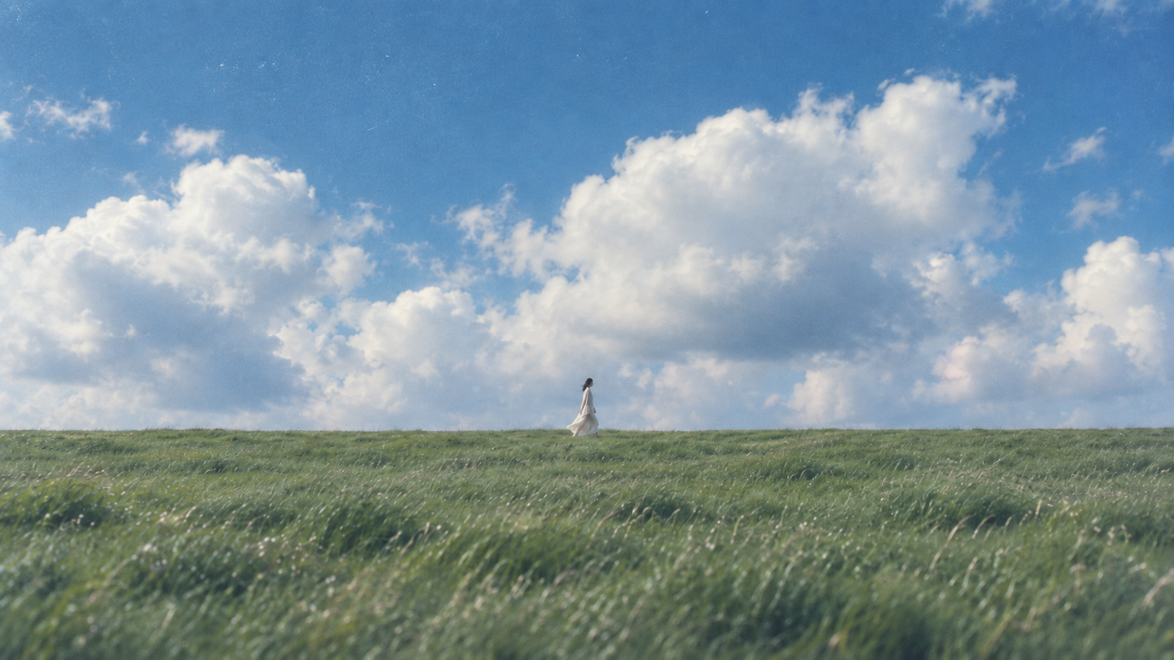 A person walking across a green field beneath a wide sky filled with clouds.